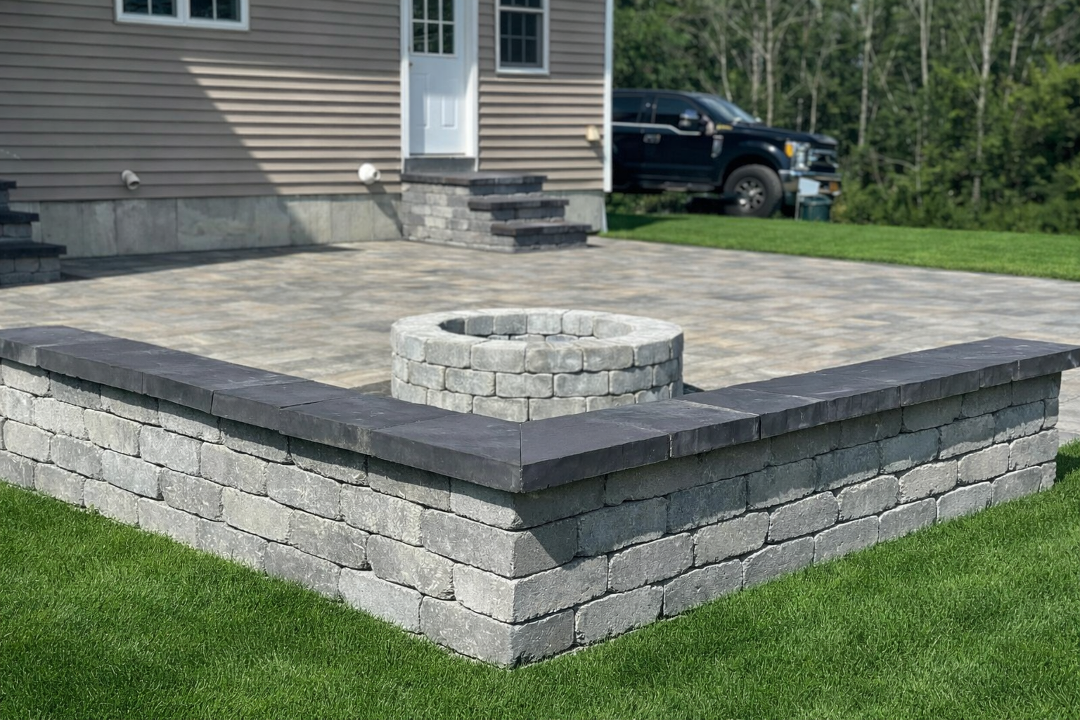 Raised paver patio with retaining wall and circular fire pit in backyard