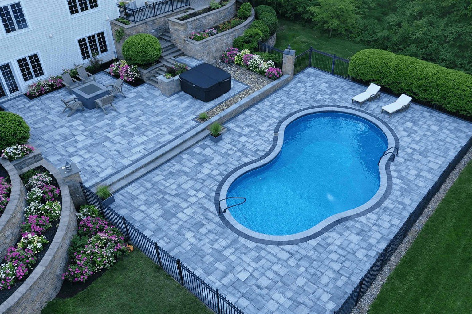 Modern backyard featuring a curved swimming pool, large paver patio, seating area, and landscaped retaining walls.