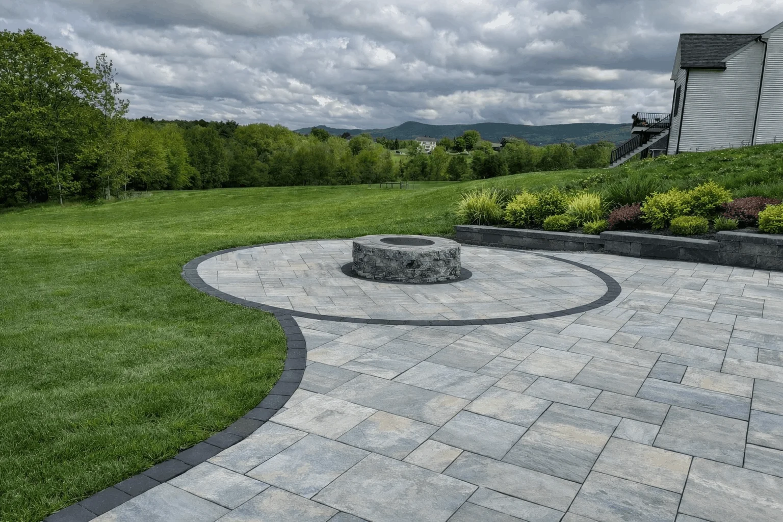 Backyard paver patio featuring round fire pit area, curved borders, and raised planting bed