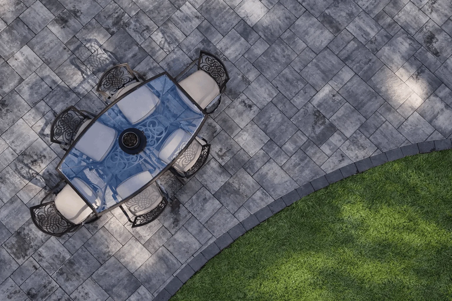 Aerial patio scene featuring outdoor dining table on patterned pavers beside lawn edge