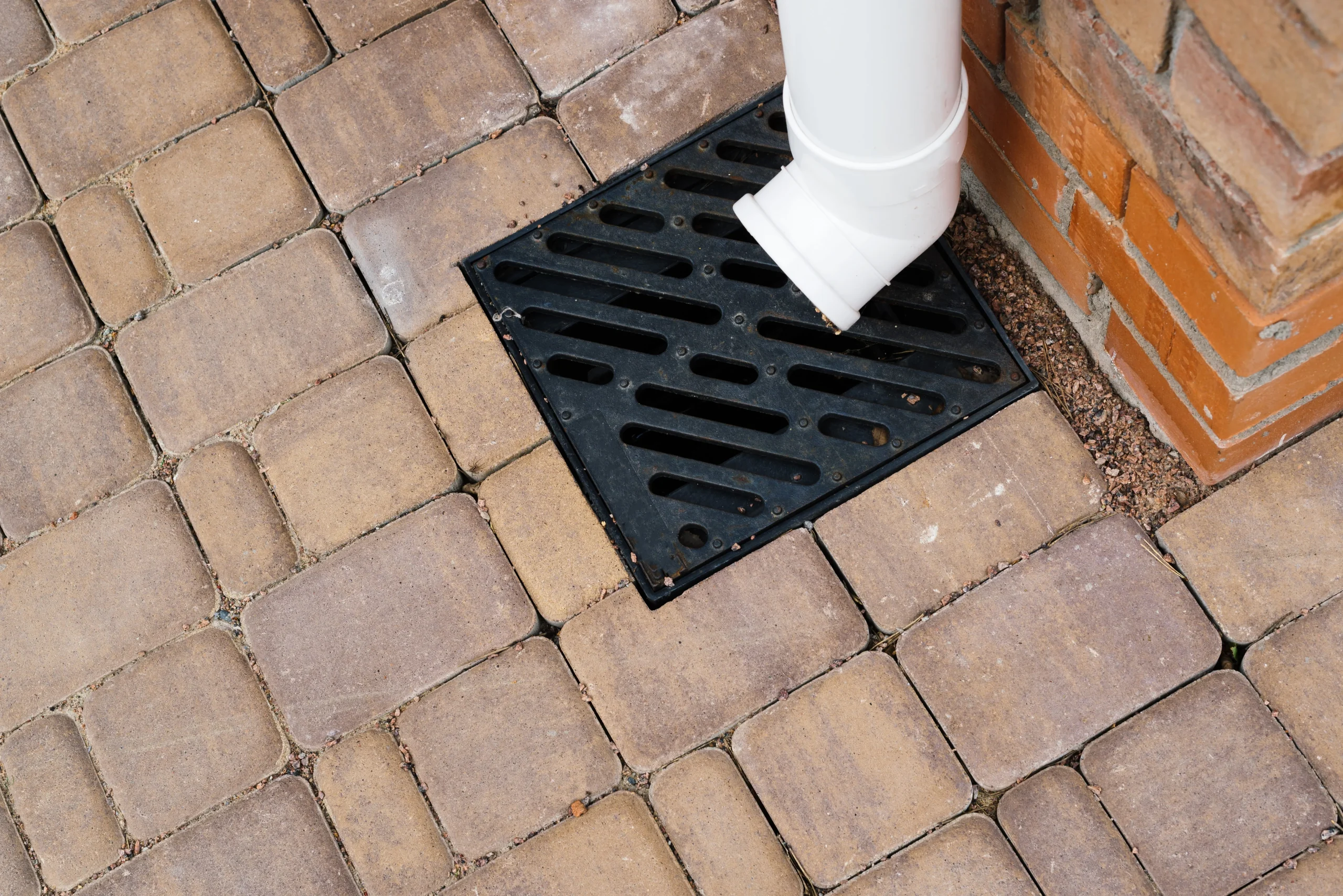House gutter downspout directing rainwater into a square outdoor drainage grate set within a stone paver surface.