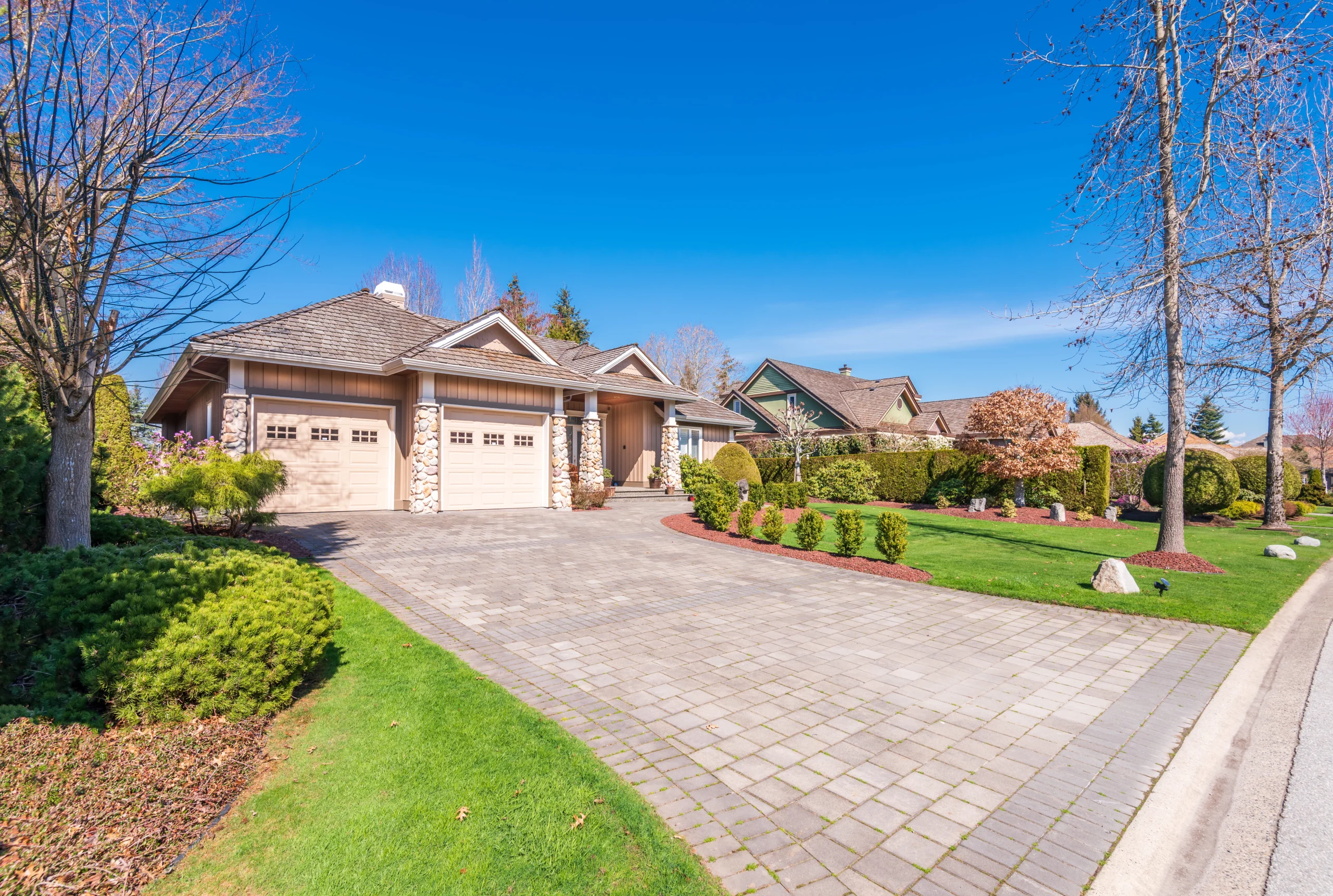 Large stone paver driveway leading to a two-car garage with landscaped lawn and trees in a suburban neighborhood.