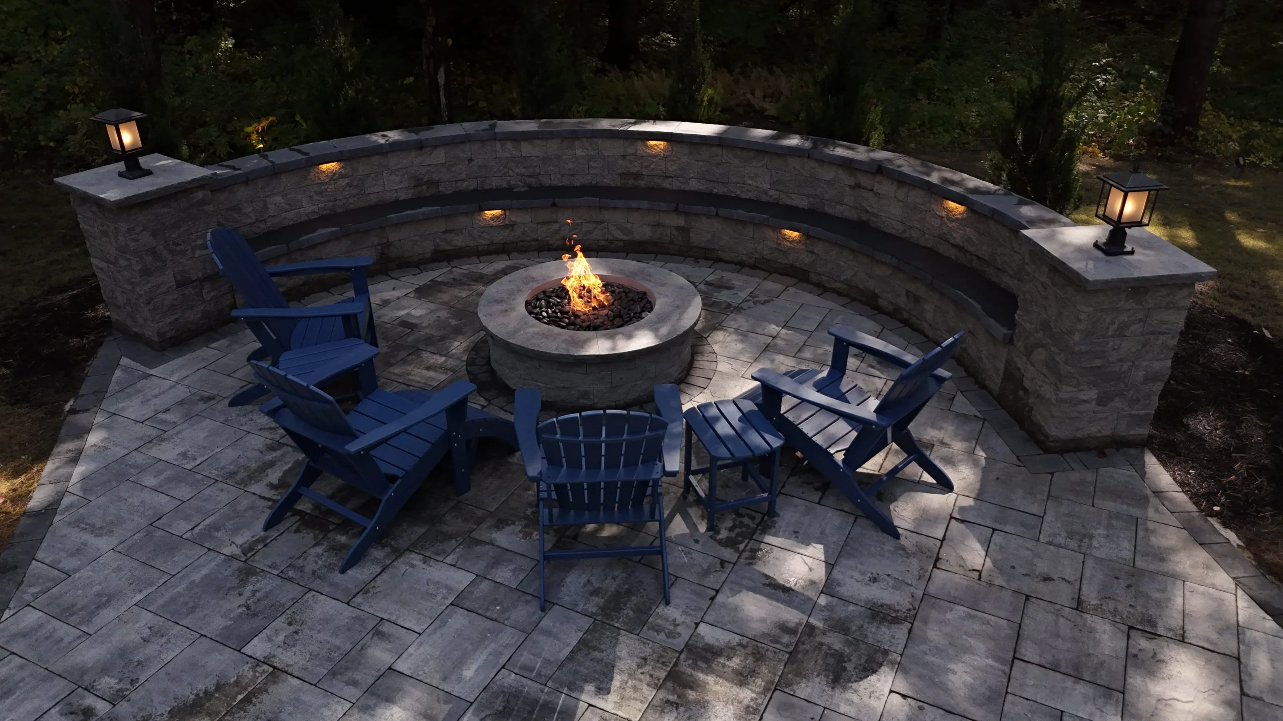 Outdoor stone patio with central fire pit, seating area, and illuminated retaining wall at night.
