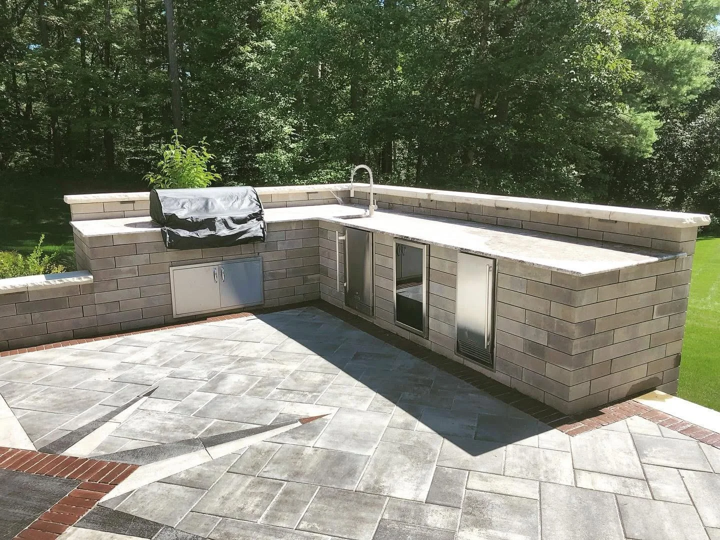 Backyard outdoor kitchen featuring a built-in grill, countertop sink, and storage cabinets on a stone patio.