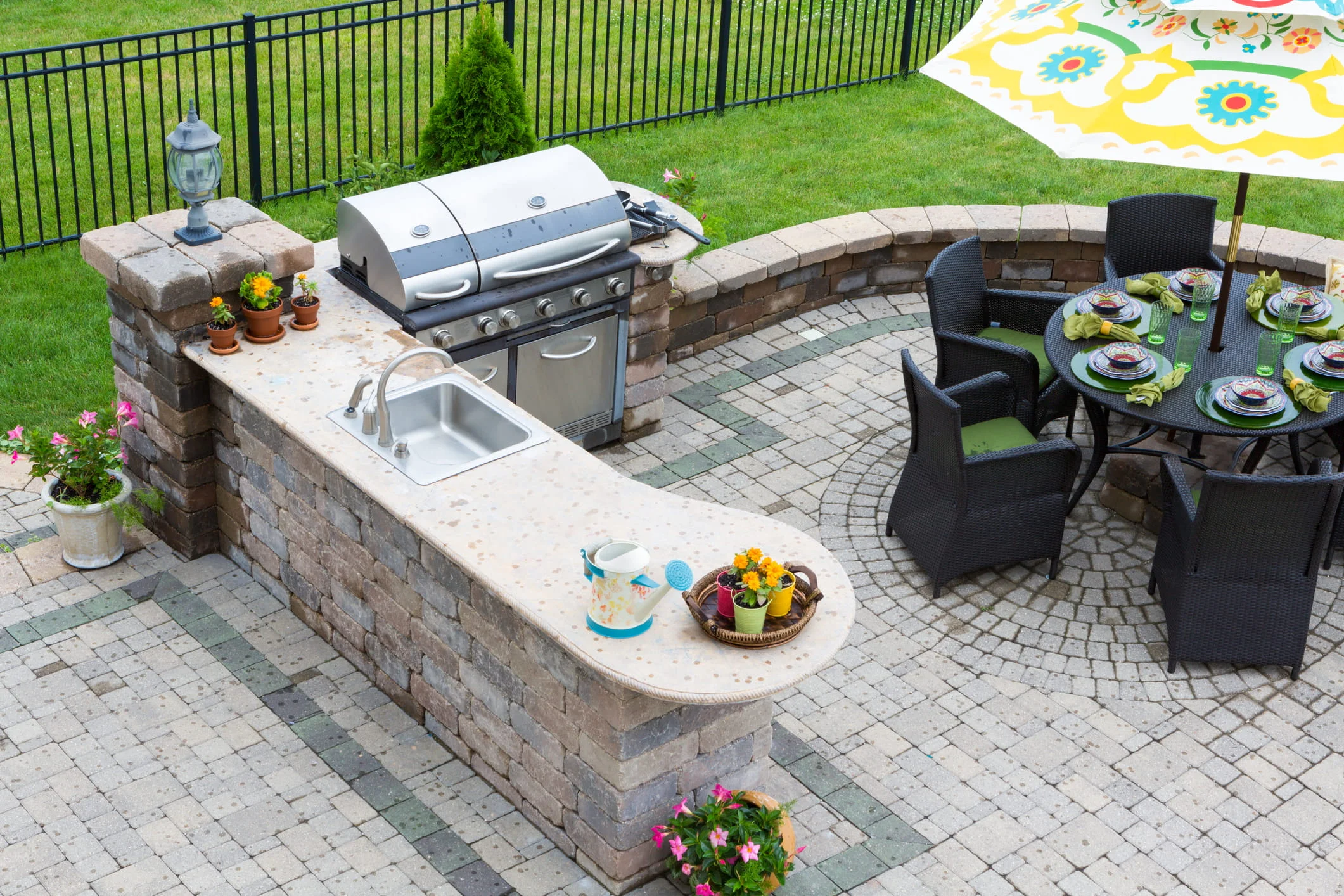 Stone outdoor kitchen with grill and sink beside a circular patio dining area with chairs and colorful umbrella.