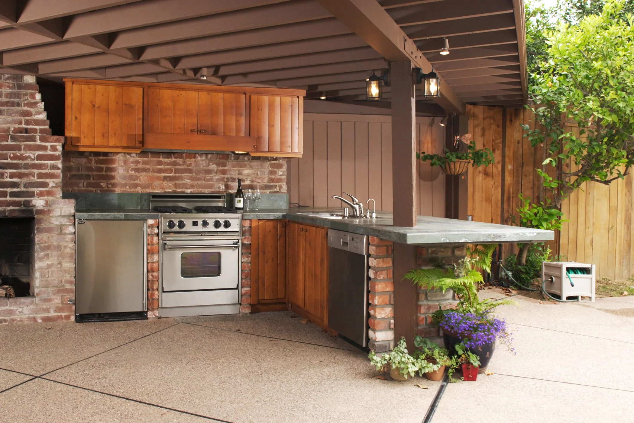 Covered patio outdoor kitchen with brick backsplash, wooden cabinets, built-in stove, sink, and countertop workspace.