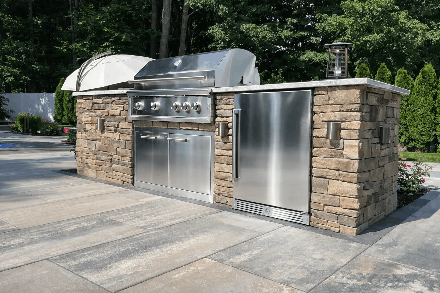 Built-in stainless steel barbecue grill and refrigerator installed in a stone outdoor kitchen on a patio.