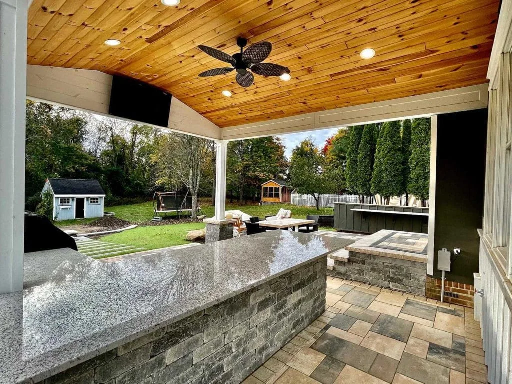 Covered outdoor kitchen patio with stone counters, ceiling fan, and backyard seating area
