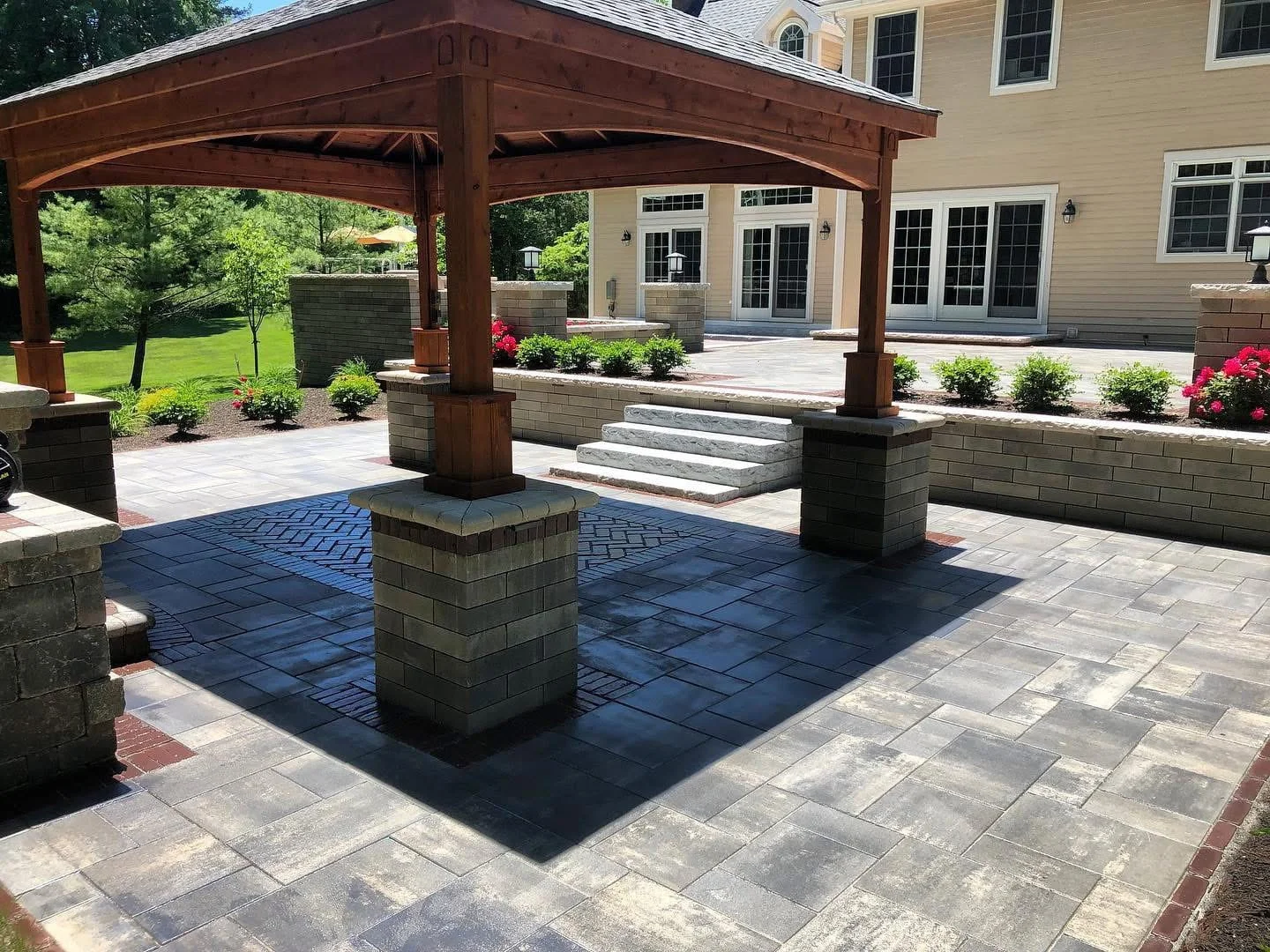 Spacious backyard patio featuring a wooden pergola, stone paver flooring, steps, and landscaped garden beds in New York.