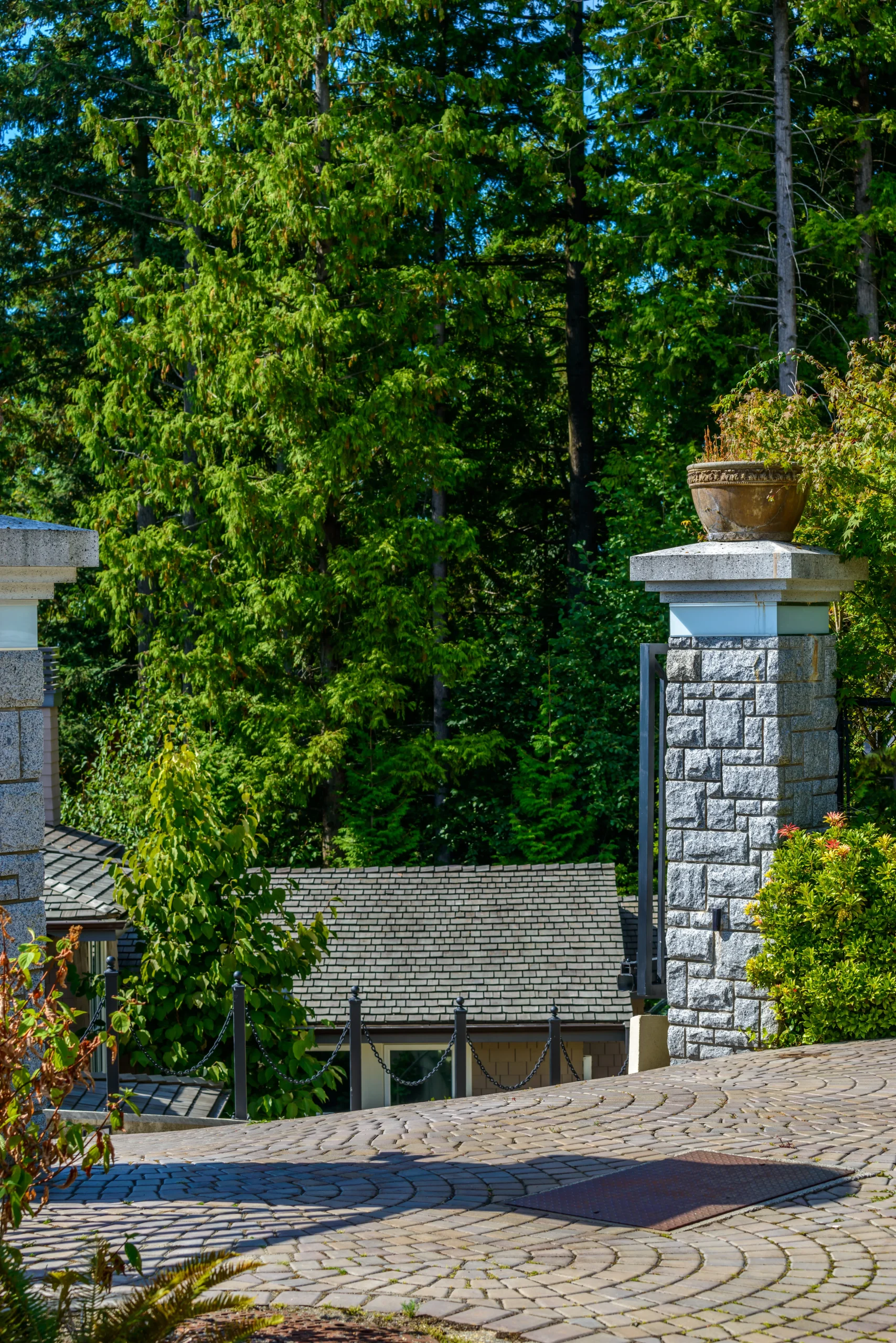 Stone pillar columns with planter pots beside a curved paver driveway surrounded by lush trees.