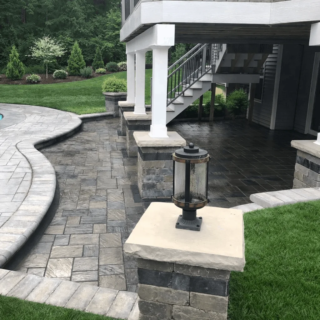 Covered patio space under a deck featuring stone pavers, stairs, lawn edging, and a pillar-mounted lantern.