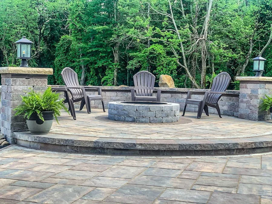 Circular stone fire pit surrounded by Adirondack chairs on a paved backyard patio.