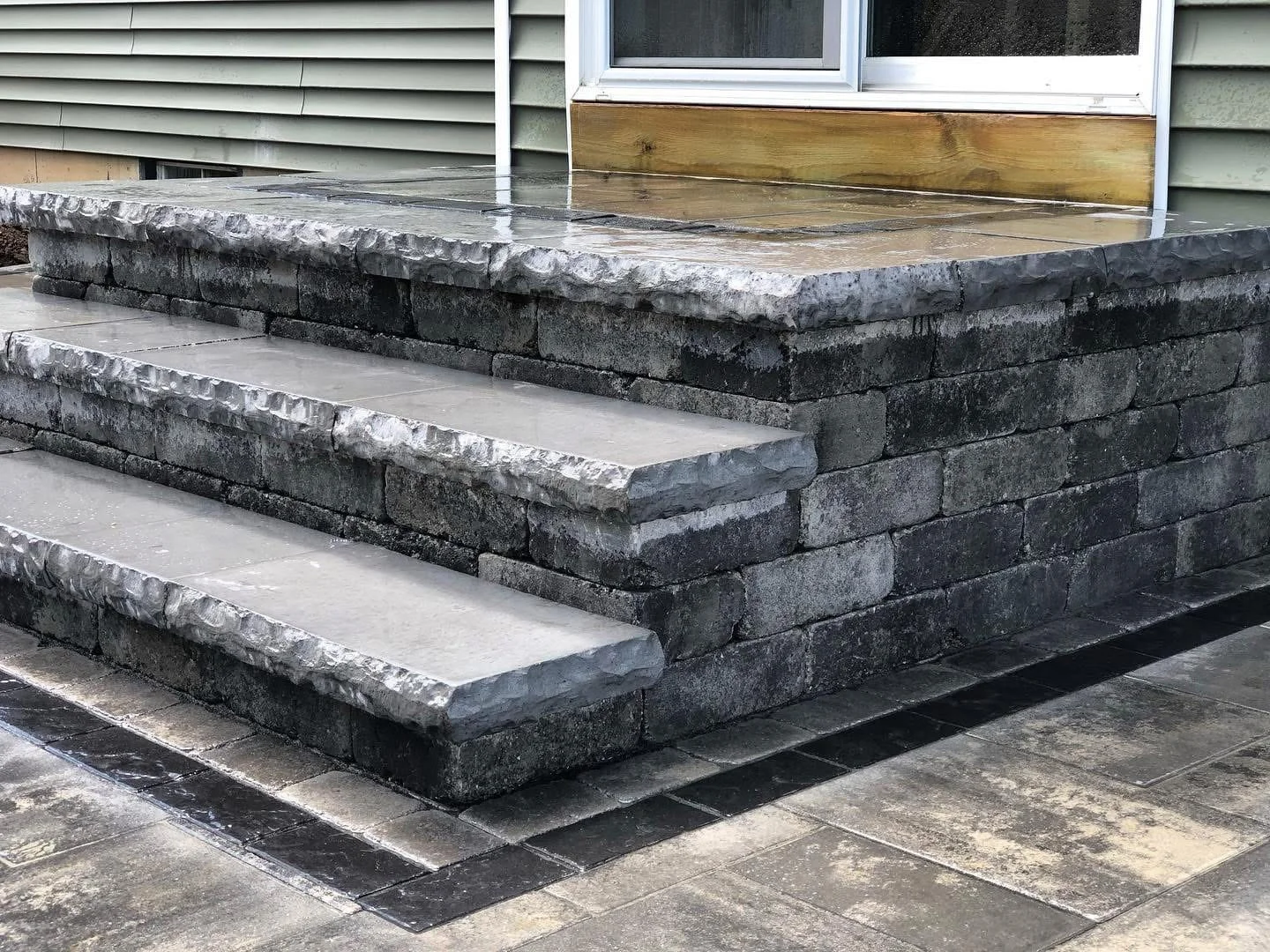 Multi-level paver patio featuring textured stone steps and a raised landing beside a house exterior.