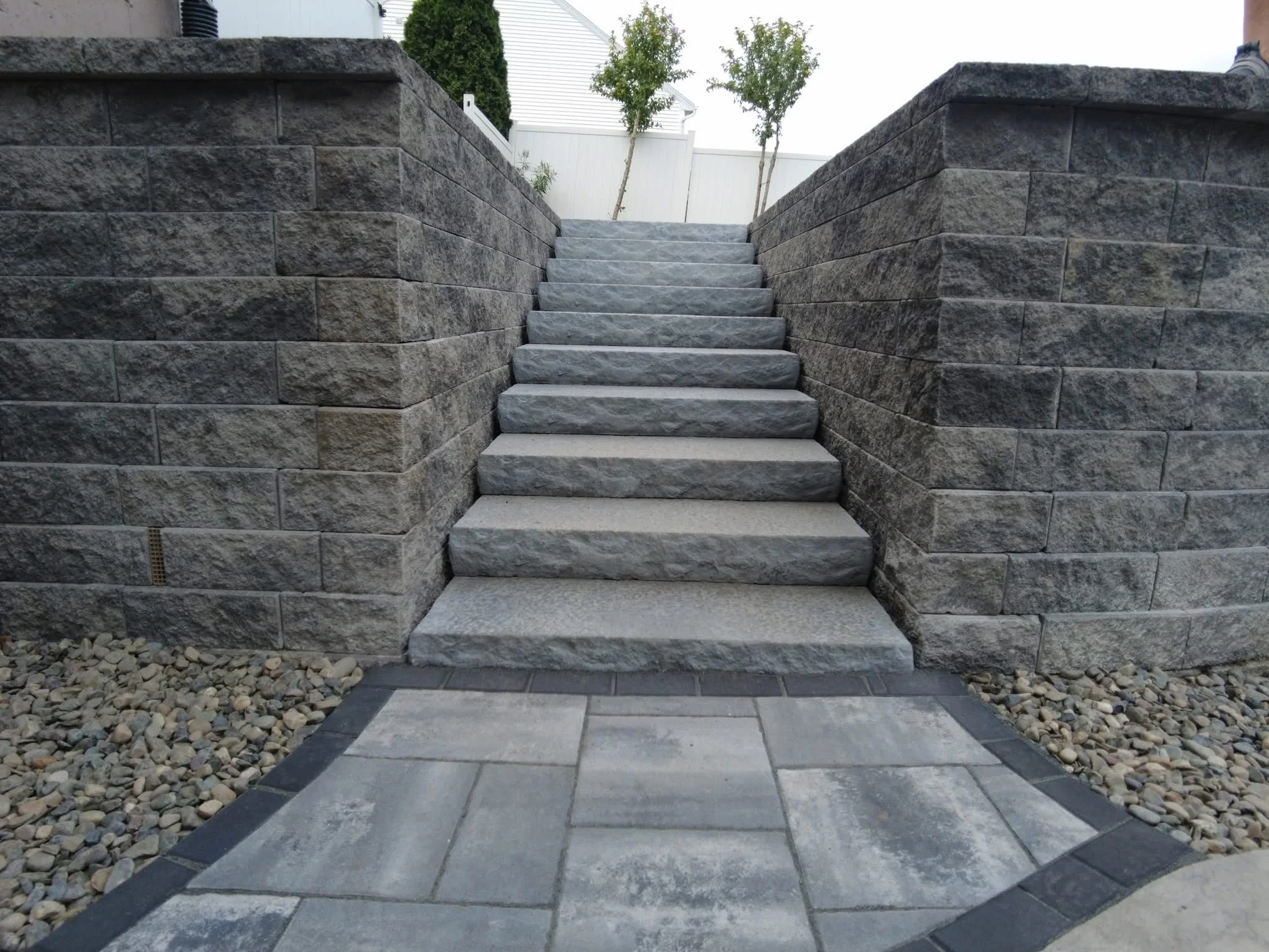 Gray stone staircase bordered by block retaining walls leading up from a paver patio walkway.