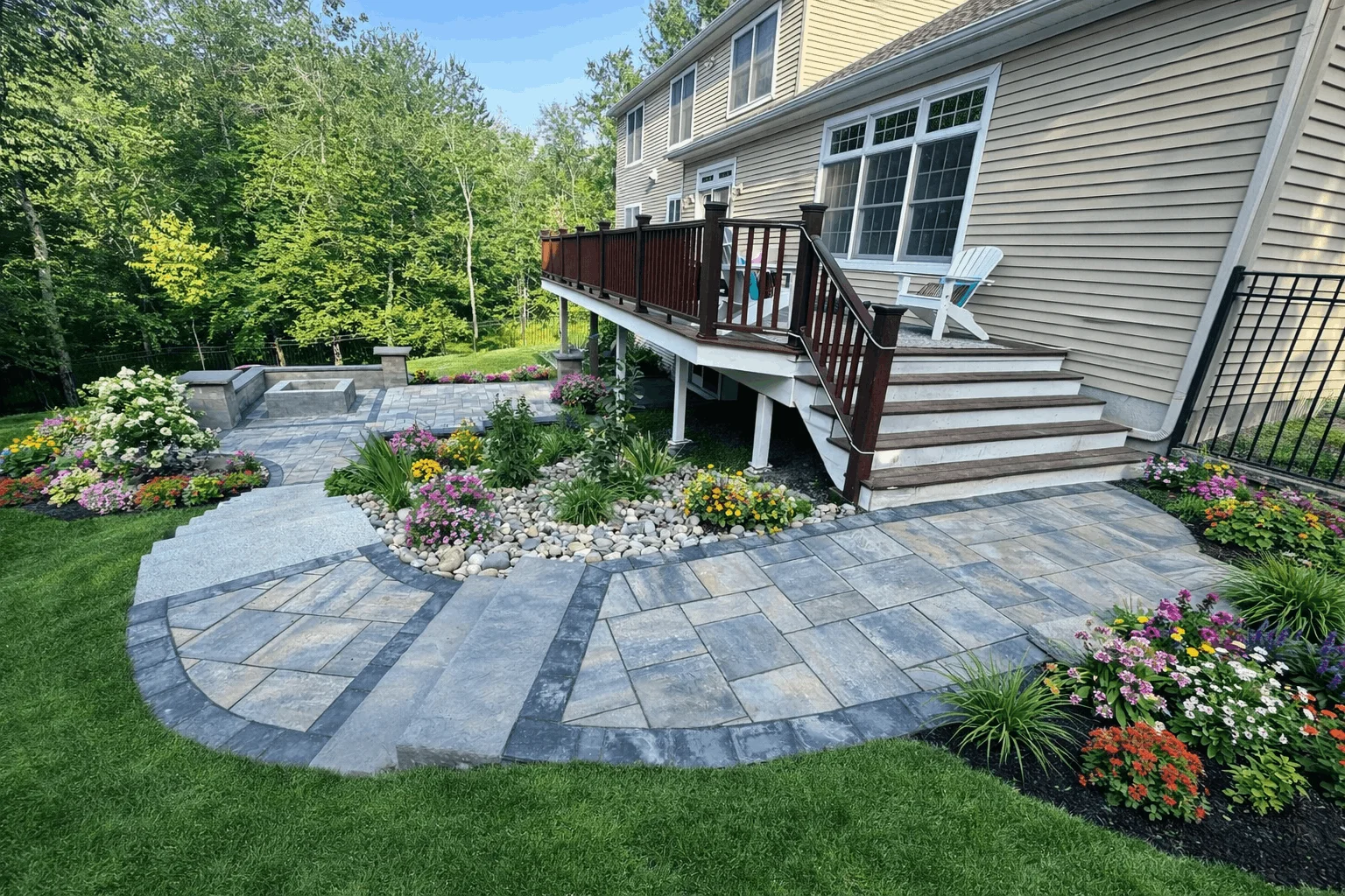 Backyard hardscape featuring stone paver patio, stairs to deck, and decorative planting beds