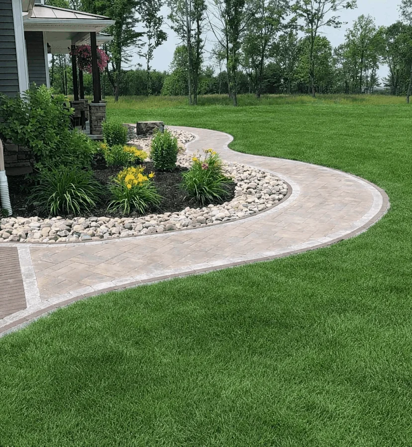 Winding backyard path made of pavers surrounded by grass, shrubs, and river rock edging